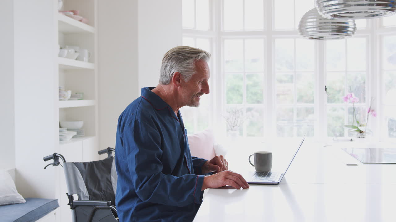 Senior Disabled Man In Wheelchair At Home Having Video Call On Laptop On Kitchen Counter