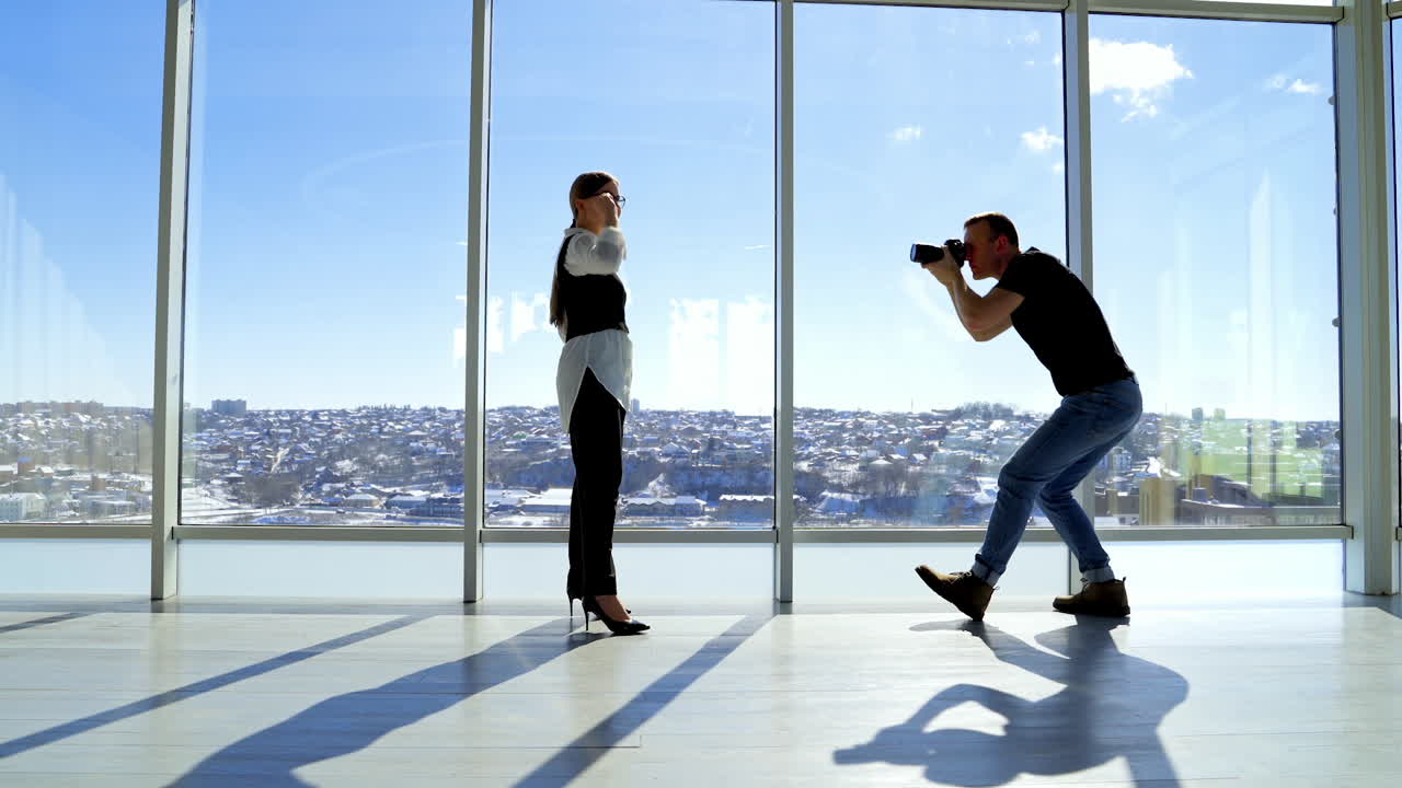 Photographer and business lady in office. Beautiful elegant woman in glasses posing on camera while standing near the window. Panoramic windows with city view.