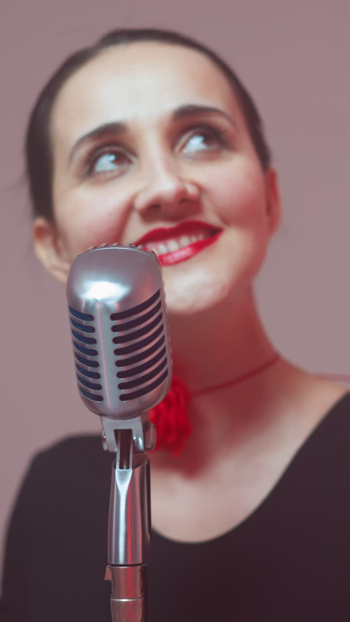 una cantante feliz con un traje negro y una cinta roja en el cuello sonríe y mira hacia arriba mientras canta en un micrófono vintage