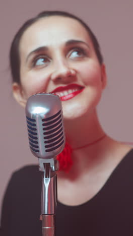 una cantante feliz con un traje negro y una cinta roja en el cuello sonríe y mira hacia arriba mientras canta en un micrófono vintage