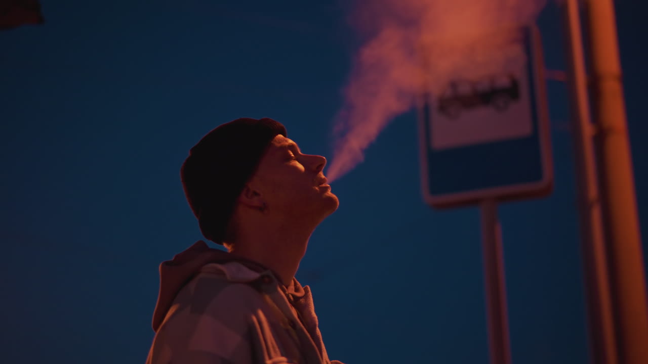 Man waiting at night bus stop, Silent night scene with man and smoke, Quiet platform with man vaping during blue dusk, Serene night scene featuring man smoking at bus shelter during twilight