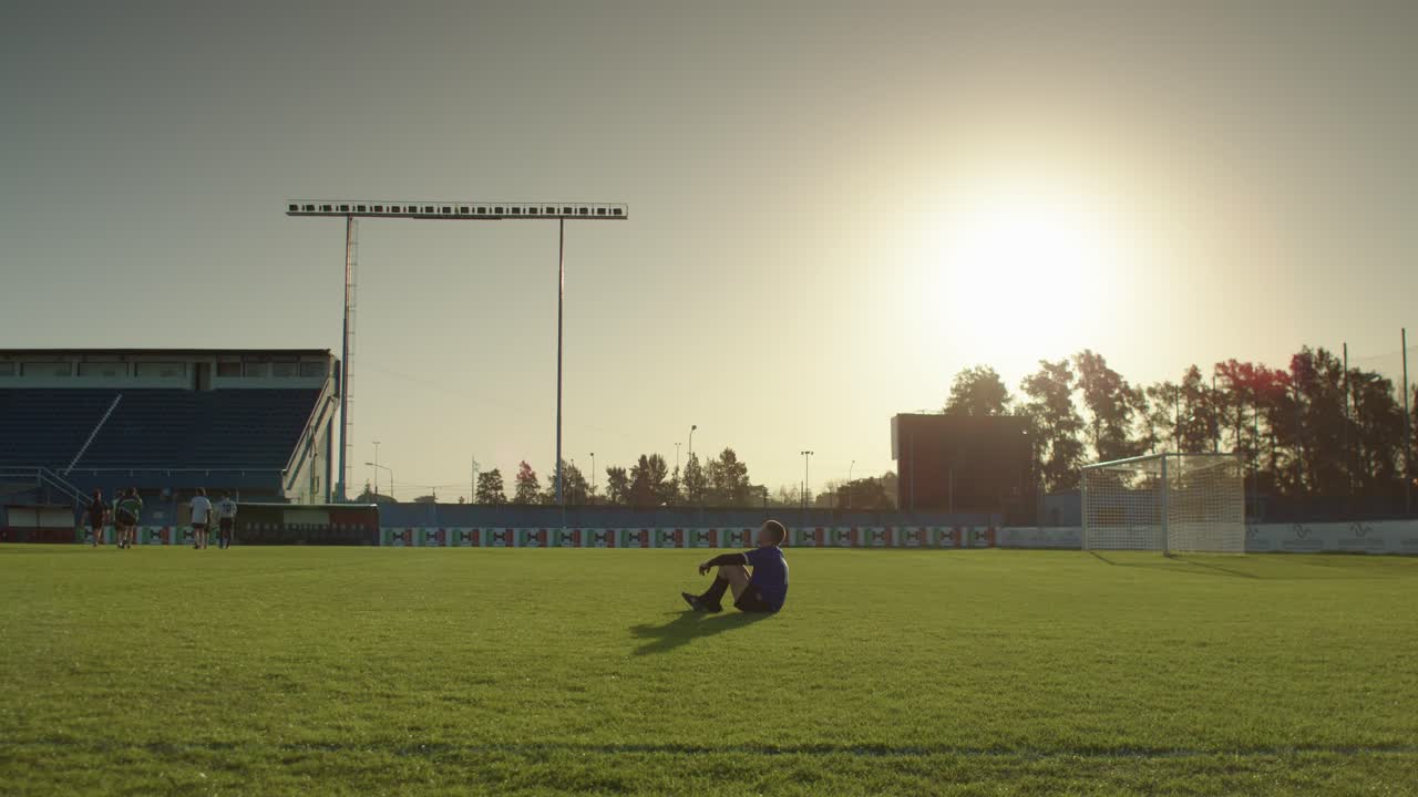 A football soccer player sits on the field resting during a sunny evening practice session
