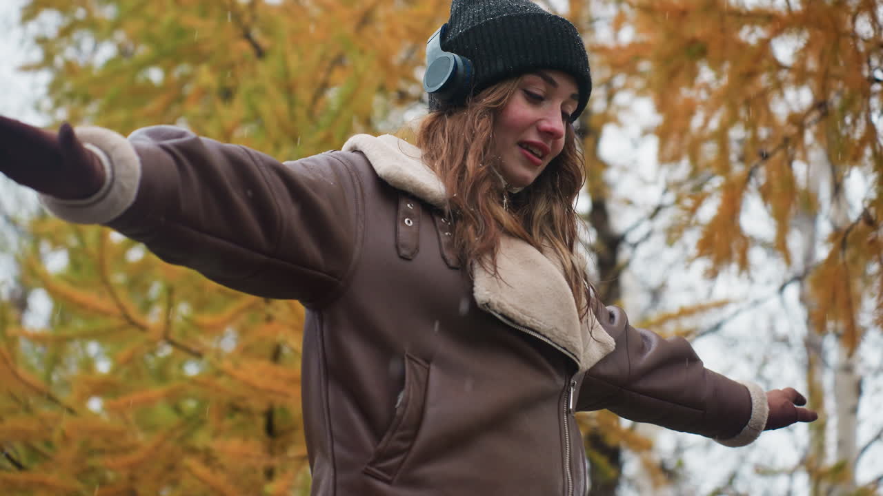 Close view of smiling female wearing black knit cap, brown shearling jacket with arms outstretched, walking joyfully outdoors on cool autumn day, enjoying nature and fresh air