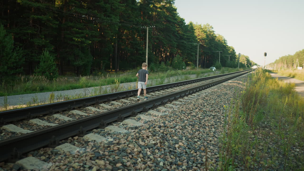 back view of child walking slowly along rail track balancing on gravel ties under warm sunlight with utility poles and parked car in distant amid forest backdrop