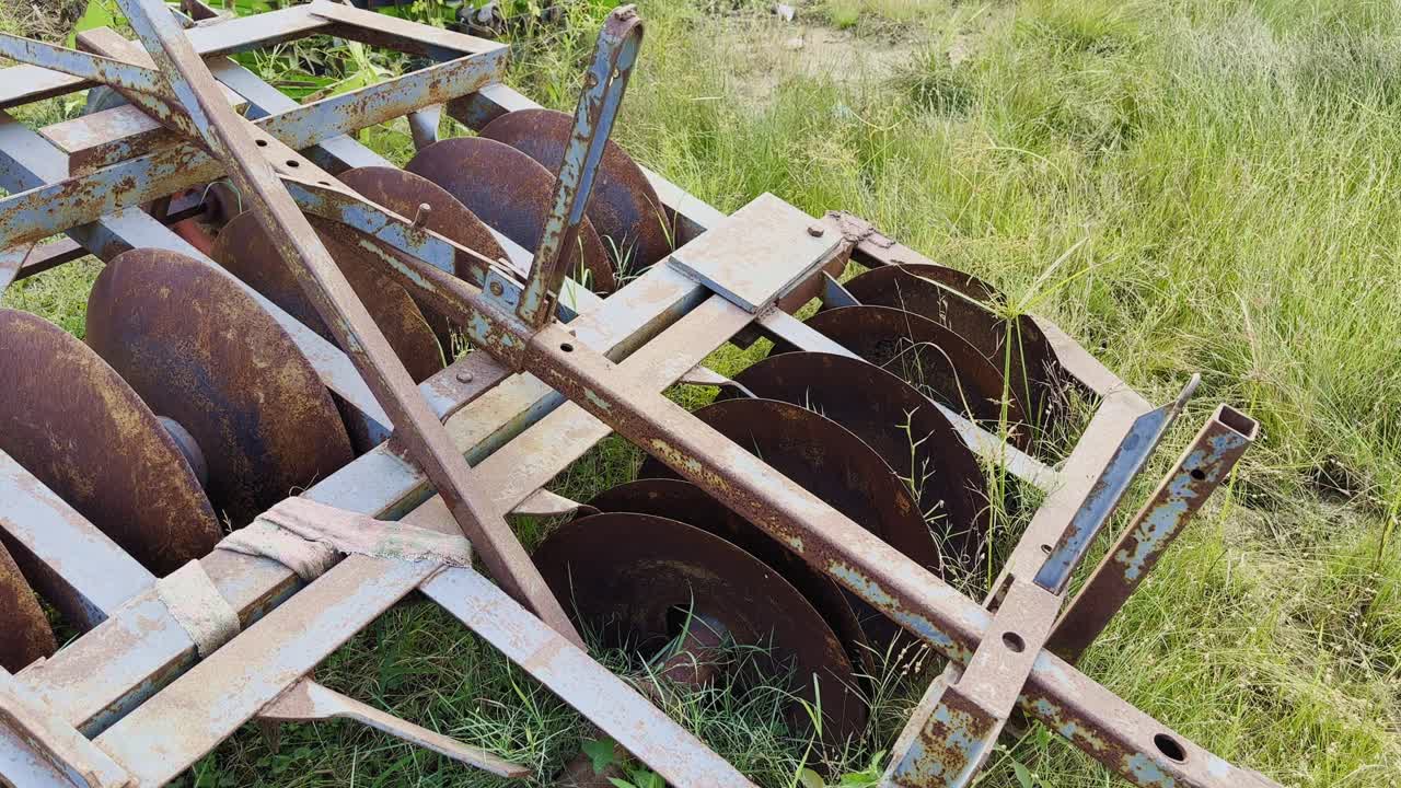 A static shot showing the circular discs of a rusty harrow resting amidst tall grass, highlighting the aging agricultural machinery left unused in the field under natural light