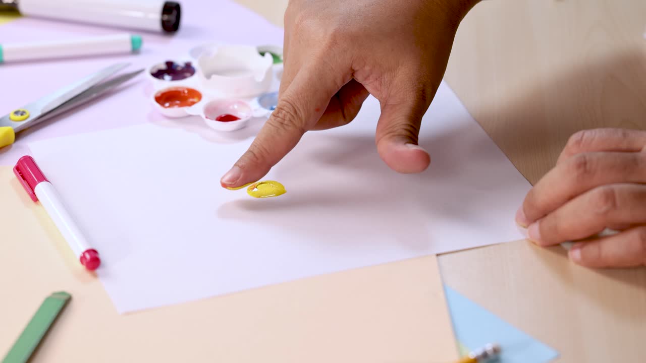 Hand dips finger in paint palette, dabs color onto white paper, bright indoor lighting