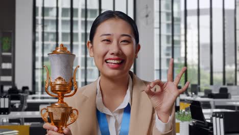 close up of asian business woman in a suit with a gold medal and trophy showing okay gesture and smiling to camera as the first winner in the office