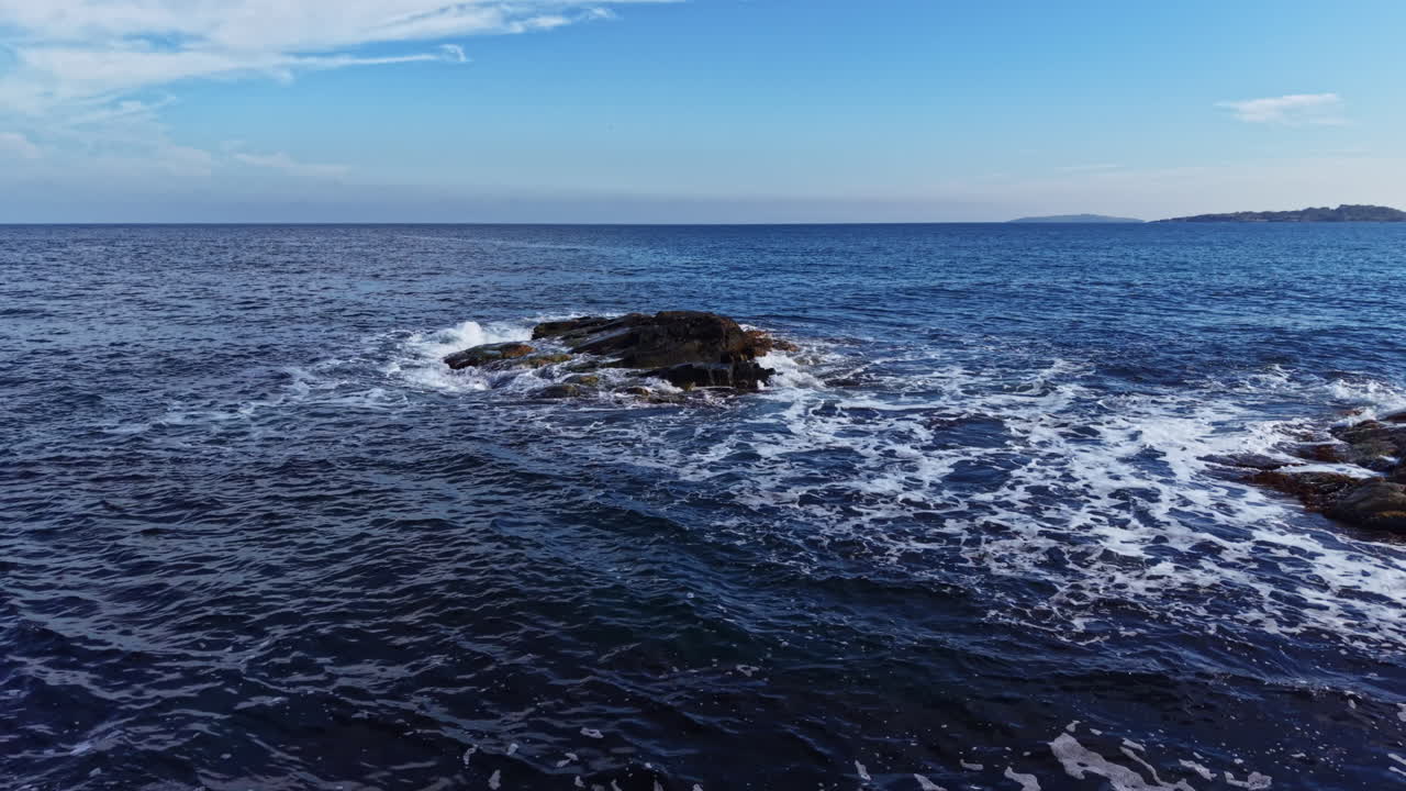 Rolige bølger slår mot steinete strandlinje under en lys blå himmel midt på dagen nær et fredelig kystlandskap.