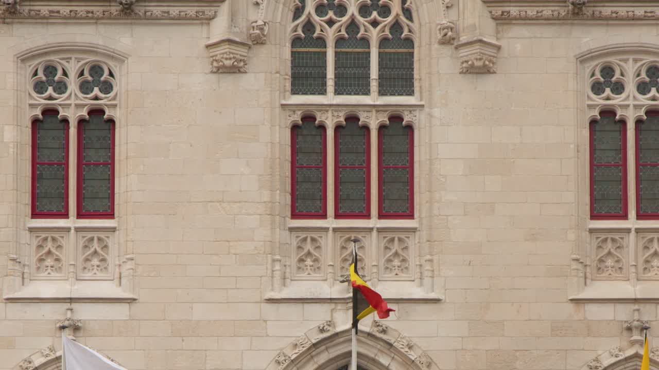Belgian flags flutter outside ornate Gothic city hall, overcast daylight, static camera, architectural detail