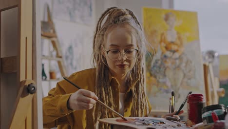 Retrato-De-Una-Joven-Bonita-Y-Elegante-Con-Gafas-Y-Rastas-Pintando-En-El-Estudio-Y-Sonriendo-Alegremente