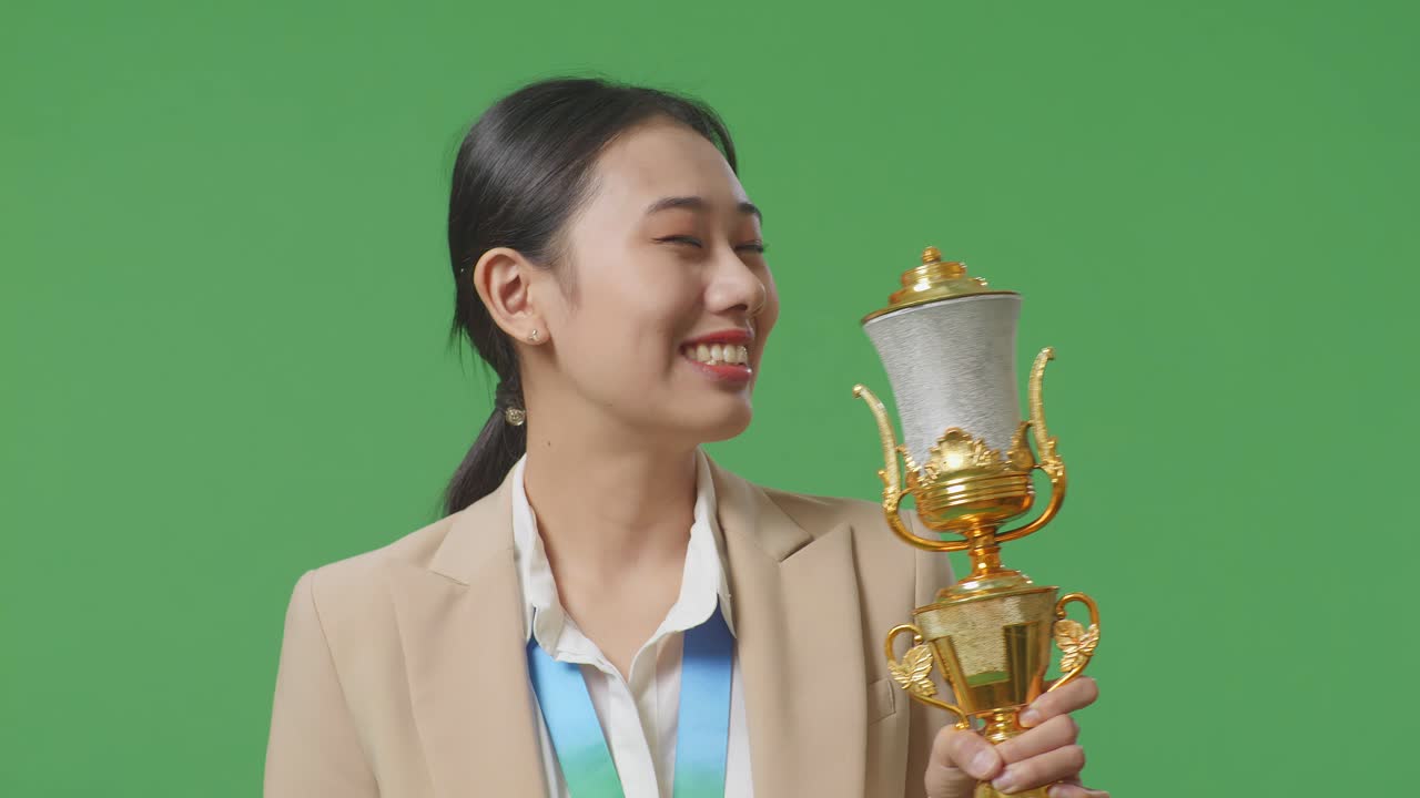 금메달을  슈트를 입은 아시아 사업가 여성이 황금 트로피를 키스하며 스튜디오의 녹색 스크린 배경에서 첫 번째 우승자로 자랑스러워합니다.