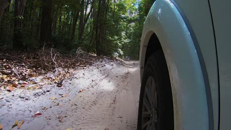 POV-Blick-Am-Vorderrad-Eines-Weißen-Allrad-Geländewagens-Vorbei-Von-Einer-Am-Auto-Montierten-Kamera,-Während-Er-Entlang-Eines-Sandigen-Binnenpfads-Durch-Den-Unheimlichen-Dschungelregenwald-Mit-Hohen-Dunklen-Bäumen-Ringsum-Hüpft