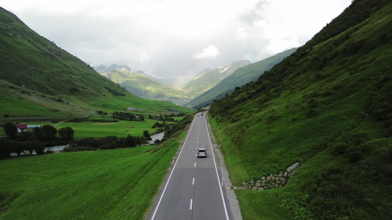 Road in Swiss Alps with small traffic car and rainbow at the sky shot from a drone