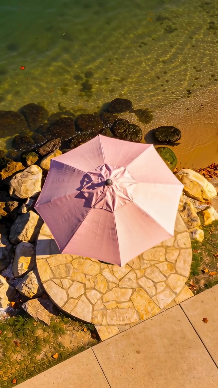 Video verticale: Drone che scende rivelando un ombrellone rosa su una piattaforma di pietra al bordo dell'acqua, mostrando rocce