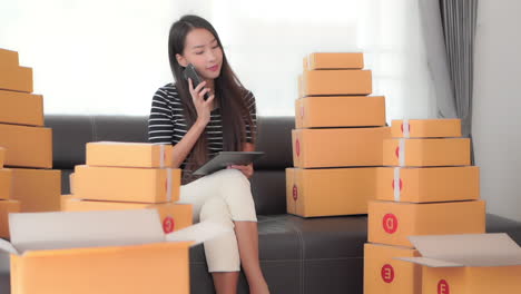 a young pretty businesswoman surrounded by packages ready for shipping checks logistics on her ipad while answering her smartphone