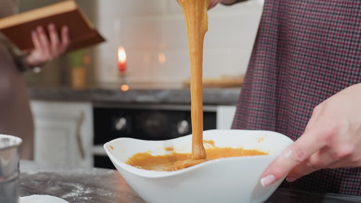 Close-up of woman holding whisk with thick caramel-colored pap stretching as it drips back into white bowl, kitchen background with blurred cookbook, candle, and flour-dusted countertop