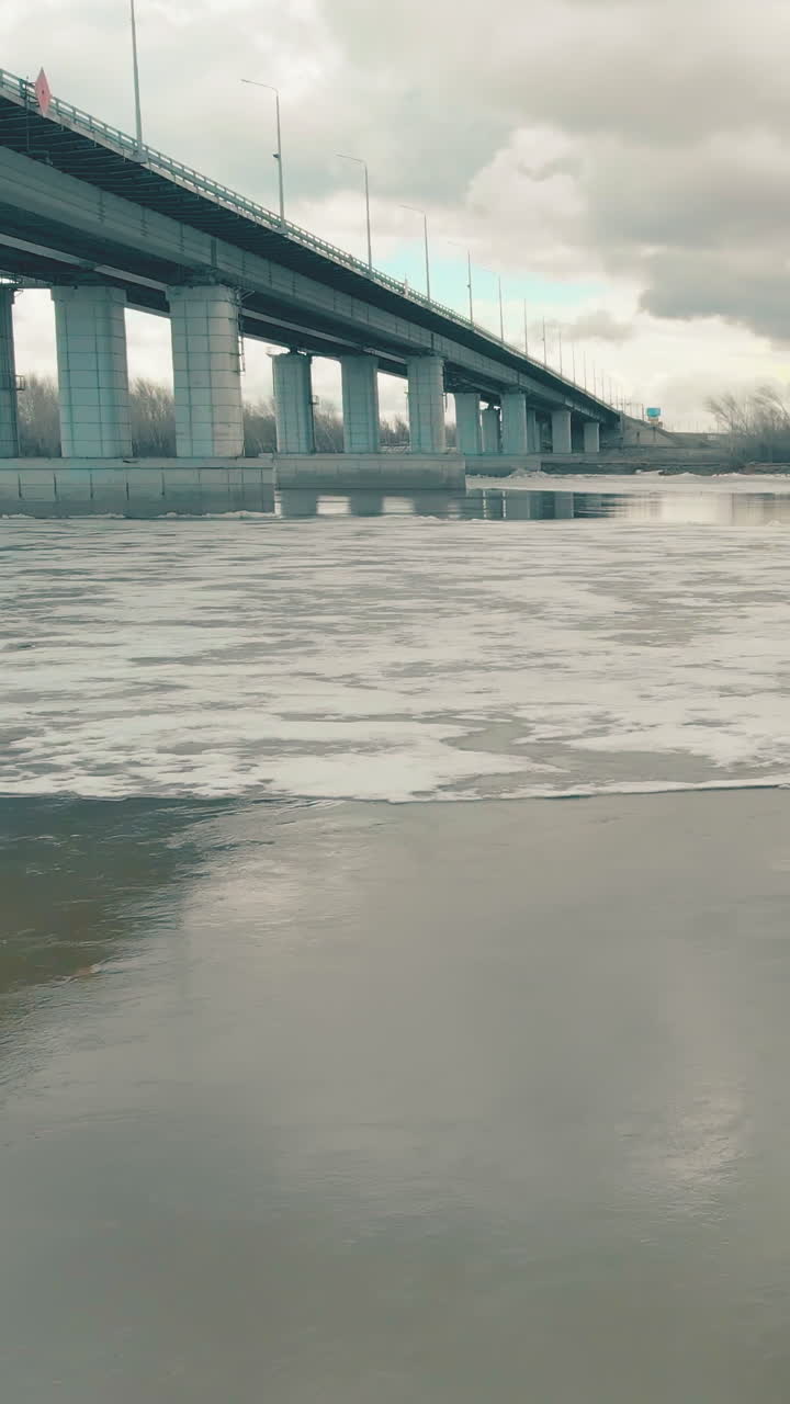 half frozen river water reflects grey bridge and clouds on patch of open water in ice on nasty winter day upper view
