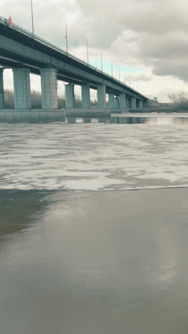 half frozen river water reflects grey bridge and clouds on patch of open water in ice on nasty winter day upper view