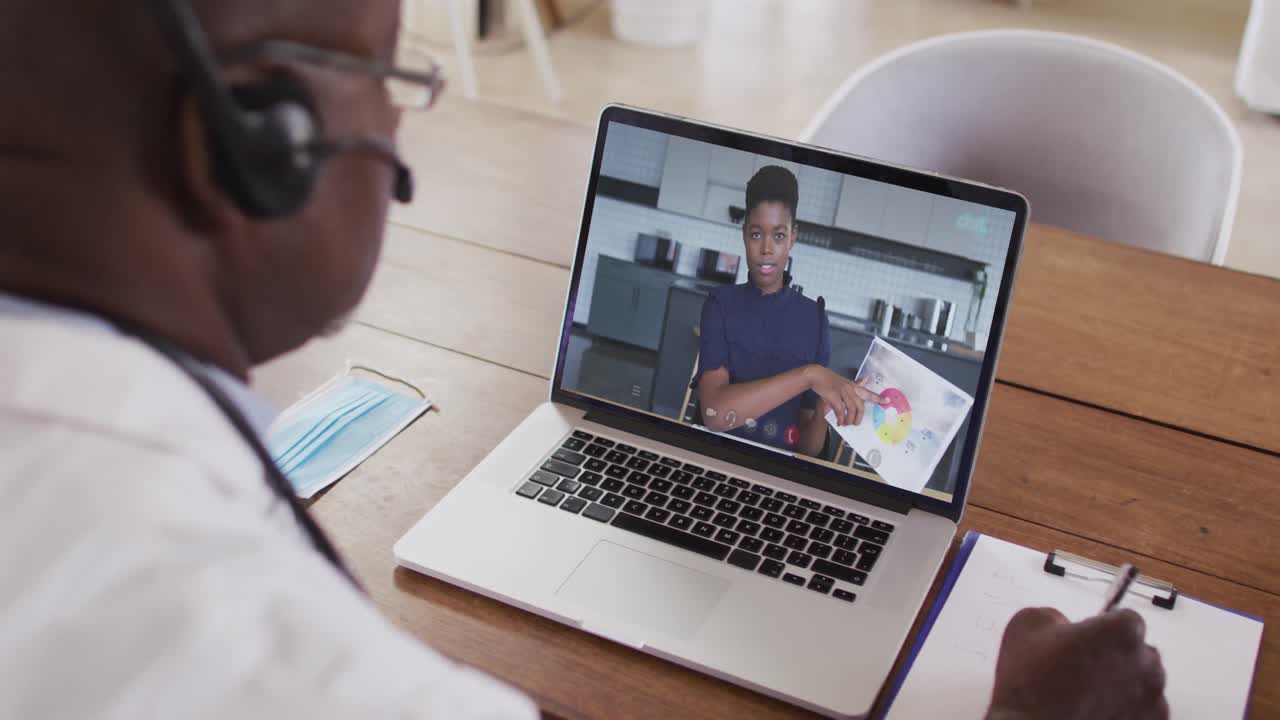 Afroamerikansk mandlig læge med headset på, der tager noter under et videoopkald på sin bærbare computer