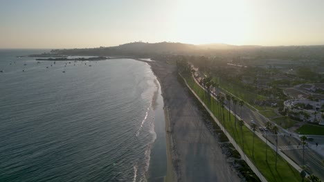 drone flying up santa barbara, ca coastline at sunset with footage of ocean waves crashing gently against the beach and people walking on the shoreline