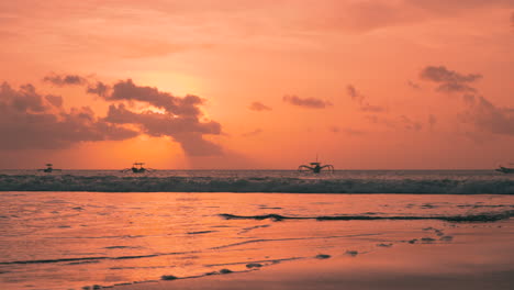 Siluetas-De-Viejos-Barcos-Con-Estabilizadores-Anclados-En-El-Mar-Al-Atardecer-De-Un-Intenso-Color-Naranja-Con-Olas-Rompiendo-Sobre-Una-Playa-De-Arena-En-Bali,-Indonesia.-Son-Varias-Embarcaciones-Con-Uno-O-Más-Flotadores-De-Soporte-Lateral.