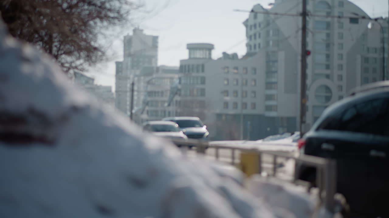 Close up snow mound by road as cars stream through lane under winter sun, modern city skyline softened in background, urban traffic mood with cold air, steady motion past curb pile