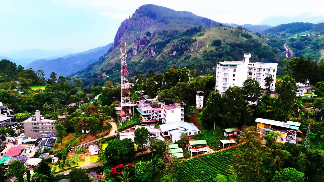 A bustling Sri Lankan mountain town stands below dramatic peaks, blending tall buildings, telecom towers, farmlands, and vibrant tropical greenery under a clear blue sky.