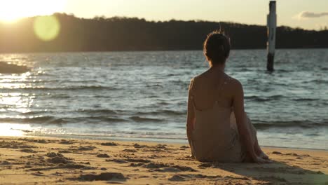 mujer joven sentada junto al agua jugando con la arena en un vestido largo durante la puesta de sol