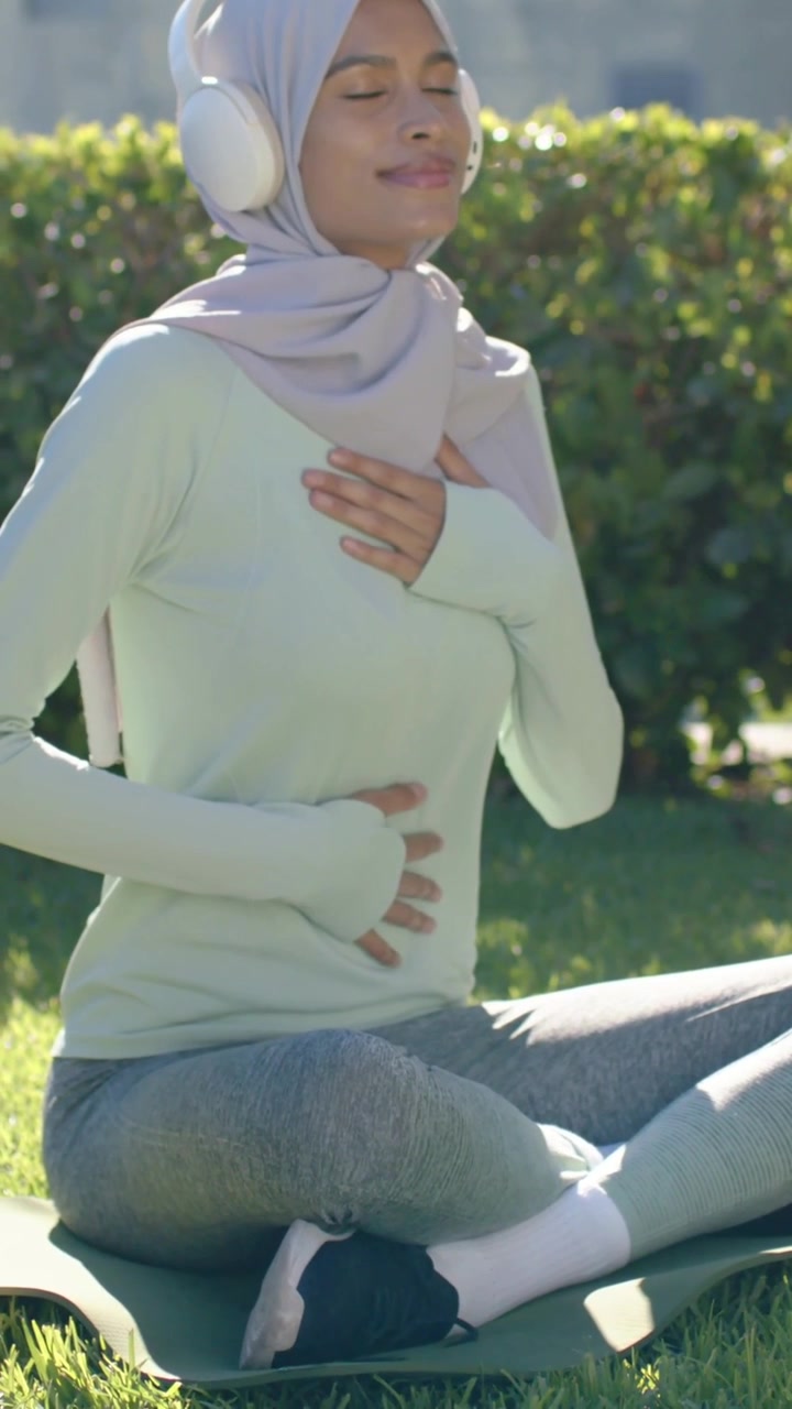Video vertikal: Memejamkan mata, wanita berhijab bernapas untuk meditasi di atas matras di taman dengan headset