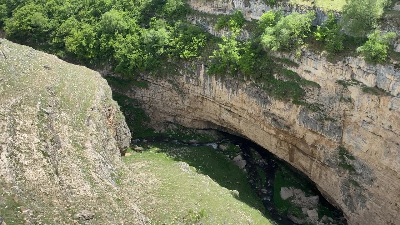 A high-angle view reveals the Karadakh Gorge in Dagestan, Russia, with a river flowing through its deep, rocky canyon and lush green vegetation on a sunny day
