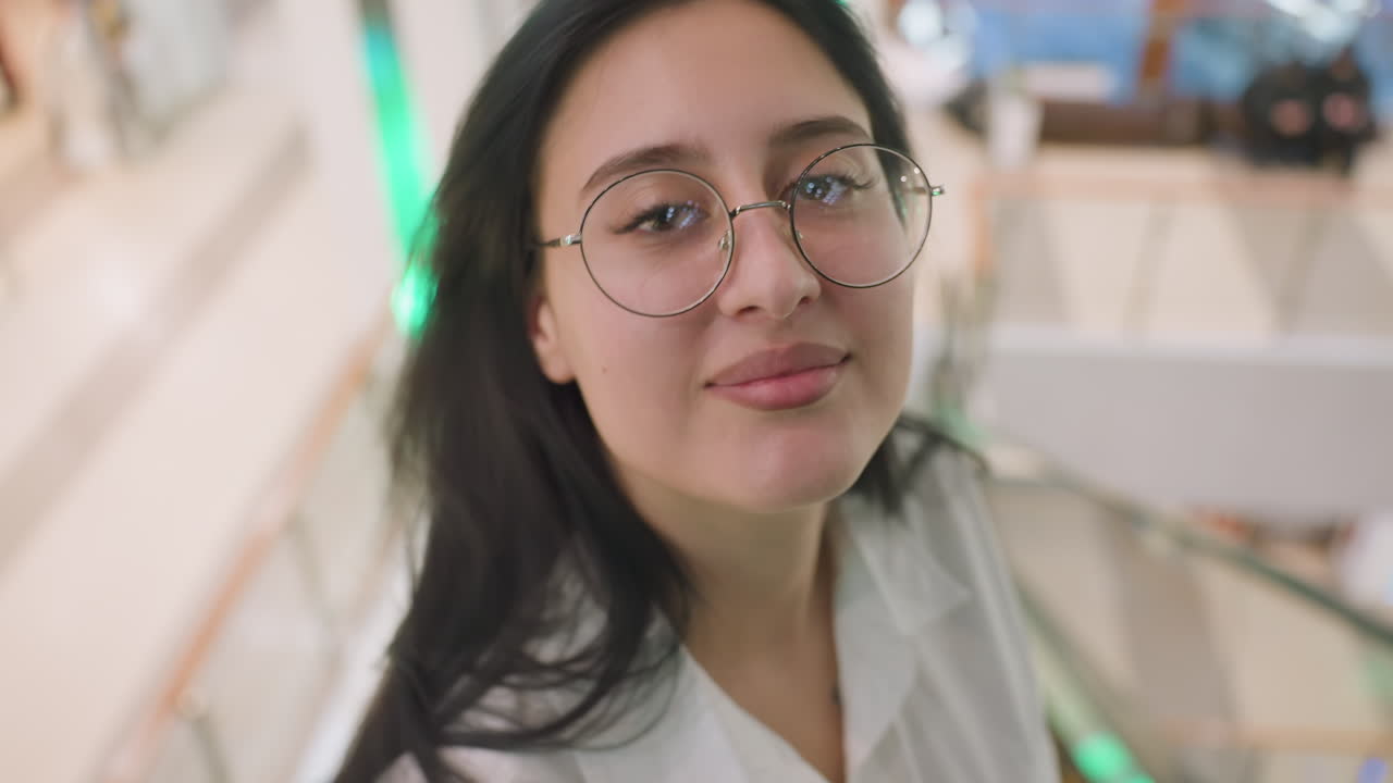 Close up of confident lady wearing glasses and white shirt brushing hair backward with hand tattoo visible, smiling slightly while posing in well-lit indoor setting with soft background blur
