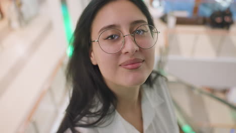 close up of confident lady wearing glasses and white shirt brushing hair backward with hand tattoo visible, smiling slightly while posing in well-lit indoor setting with soft background blur