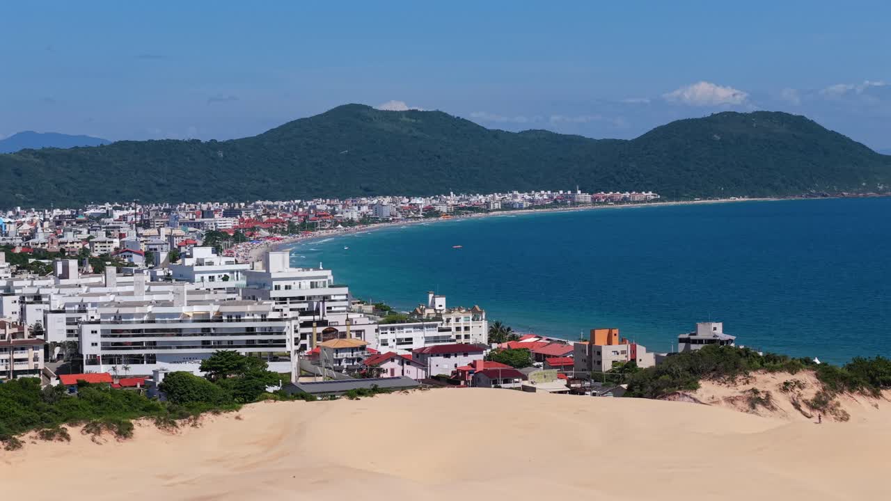 Beyaz kum tepelerinin üzerinden sakin bir koy ve yerleşim alanına doğru ileri havadan süzülüş. Güneşli bir günde Florianópolis'teki Praia do Santinho'da yeşil tepeler manzarayı çevreliyor.