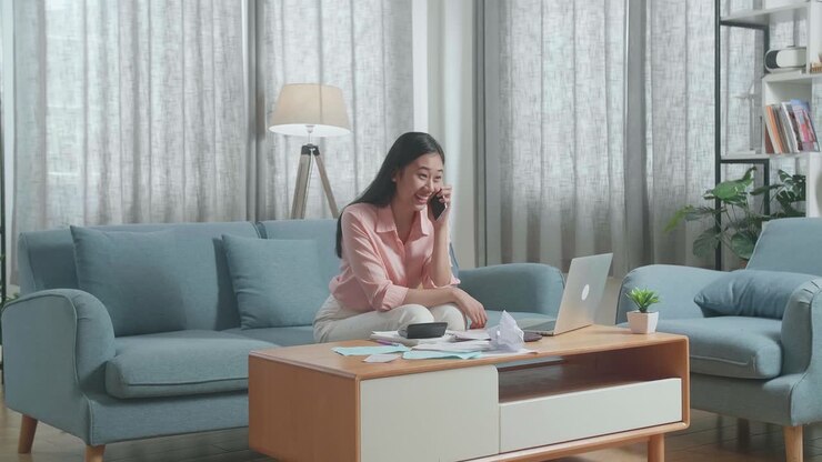 eine asiatische frau mit einem laptop, die den anruf entgegnet und dann glücklich ist, während sie auf die rechnung in der hand schaut