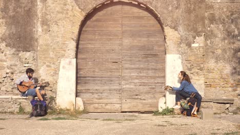 pareja feliz de mochileros turistas tocando la guitarra y cantando sentados cerca de la puerta de madera de un edificio medieval en el parco degli acquedotti ruinas del parque en roma al amanecer con guía de mapa cámara constante en cámara lenta