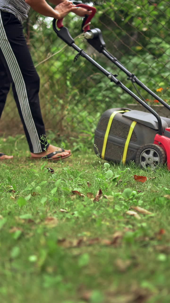 Woman working with electic mower machine in the garden. Lawn mower cutting green grass outdoors. Vertical video