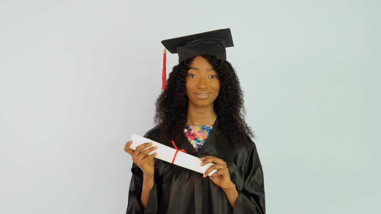 una joven afroamericana con una bata negra y un sombrero de maestro se pone erguida con el diploma de graduación en la mano y mira a la cámara. retrato de media altura