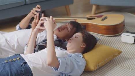 asian teen couple enjoy playing game on smartphone together while lying on carpet on the floor at home