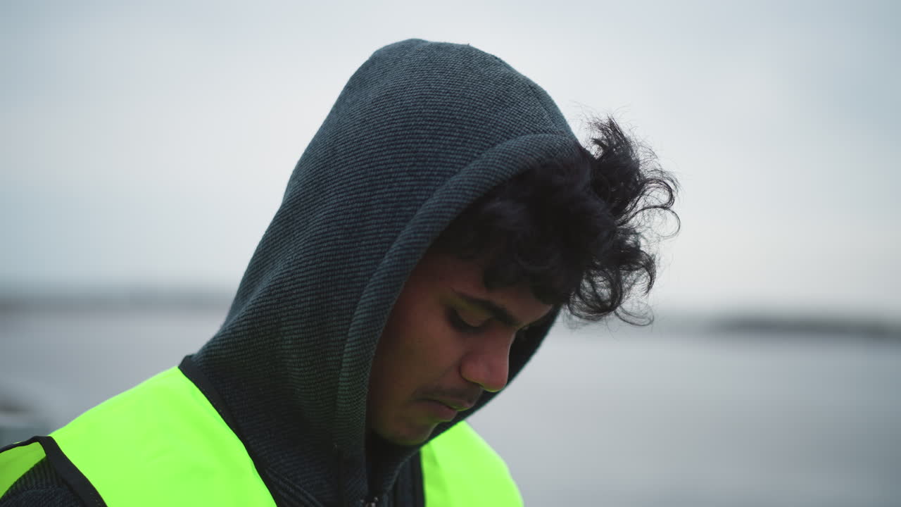 Young man with curly hair wearing dark hoodie and bright reflective safety vest standing outdoors near waterfront on cold overcast day, looking down with focused and thoughtful expression during work