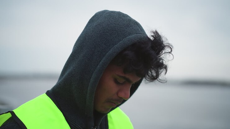 Young man with curly hair wearing dark hoodie and bright reflective safety vest standing outdoors near waterfront on cold overcast day, looking down with focused and thoughtful expression during work