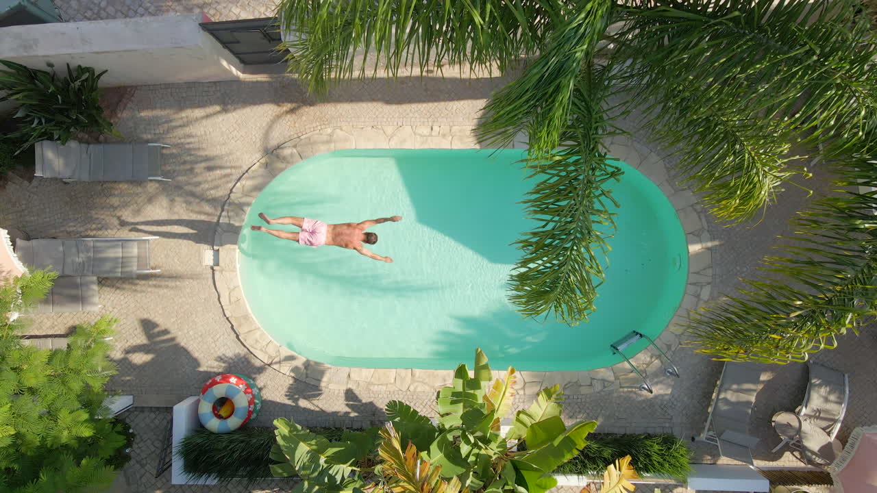 Aerial view of a man mid air as he jumps into a turquoise swimming pool surrounded by palm trees and sun loungers at a tropical villa
