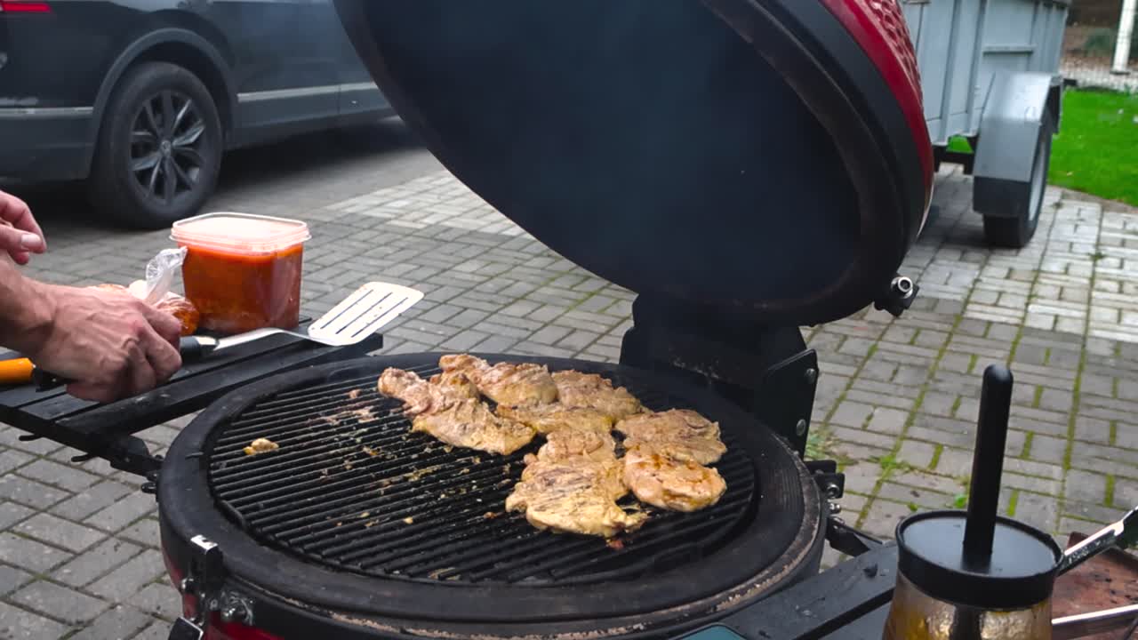 Hand with a metal spatula moving golden brown chicken meat on a kamado egg grill or barbecue outdoors during summer day in a garden. Slow motion movement while embers and smoke are flying from grill