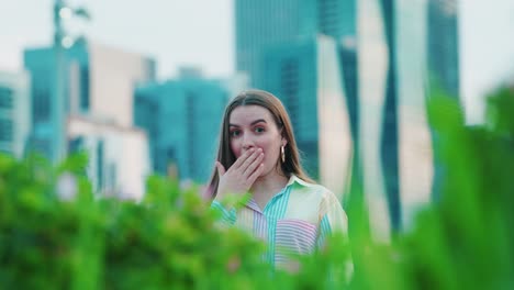 Hermosa-Chica-Caucásica-Sorprendida-Cubriendo-La-Boca-Con-La-Mano,-Luz-Del-Día,-Afuera-En-Chicago-Con-Rascacielos-En-El-Fondo-Y-Arbustos-Verdes-En-Primer-Plano