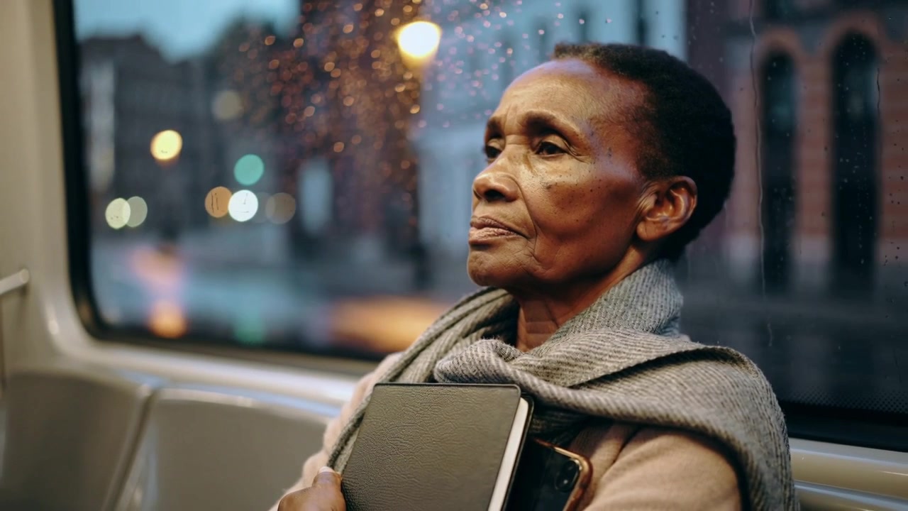 Sitting senior woman holding notebook gazing and thinking while transit moves at dusk wearing scarf
