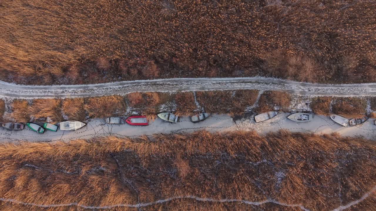 Top-down luftfoto af små fiskebåde, der ligger langs en smal, frossen kanal omgivet af tørre siv og vintergræsland