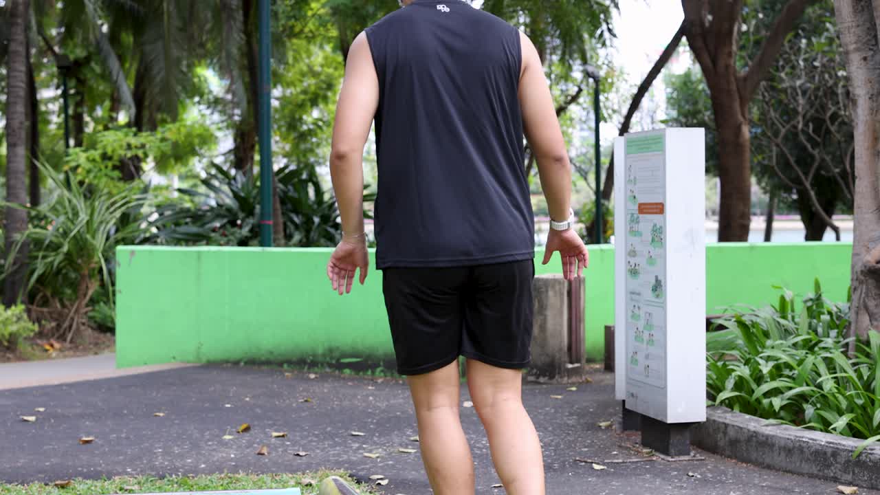 Seorang pria Asia mengenakan pakaian olahraga berjalan di atas balok keseimbangan di taman yang cerah.