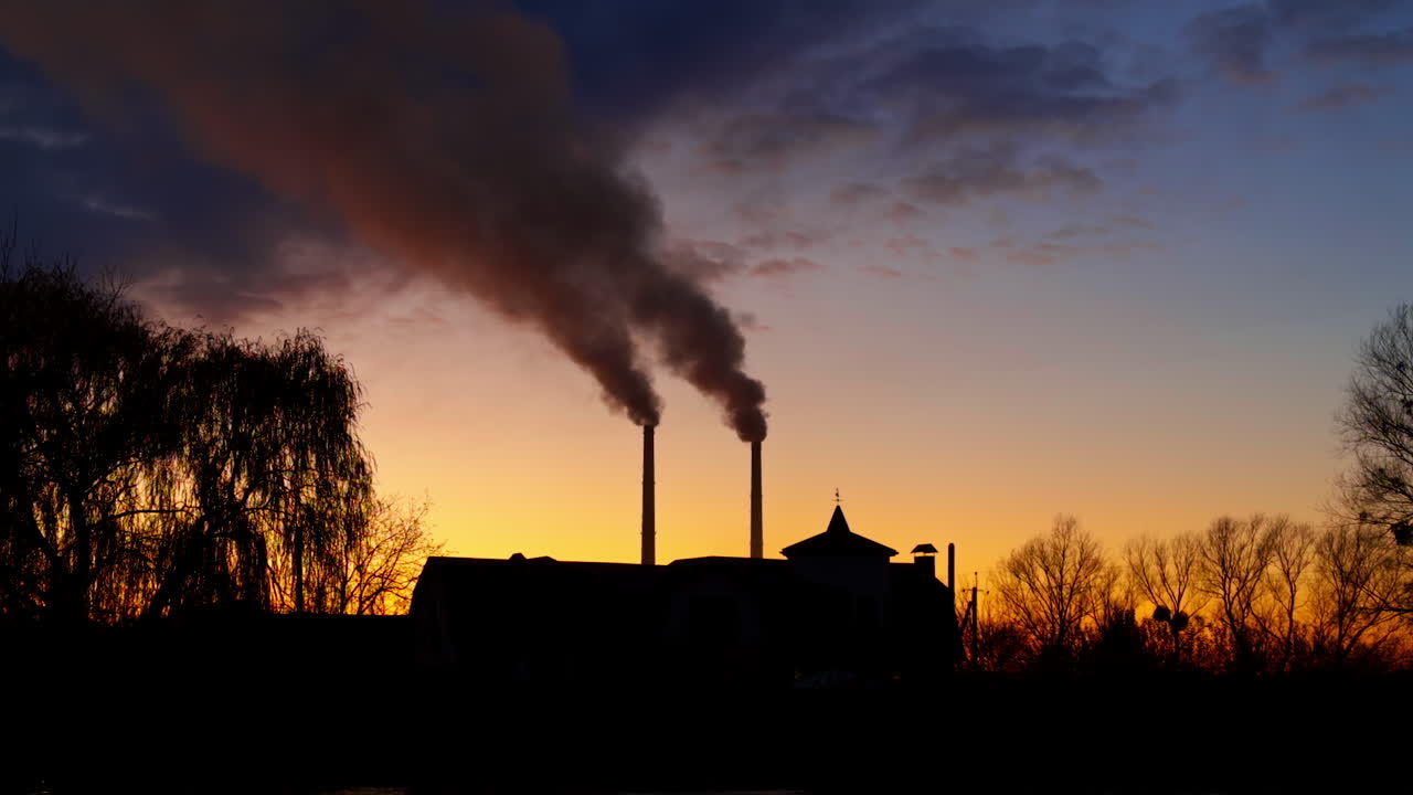 Beautiful autumn sky at sunset with silhouettes of a building, trees and plant pipes. Dark smoke goes up from the pipes.