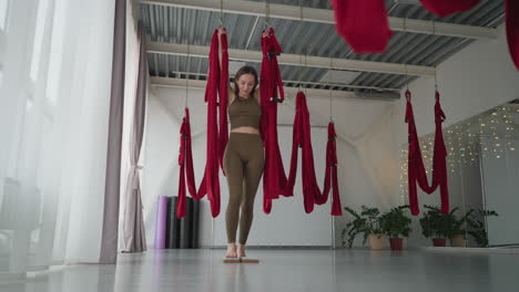 cheerful fitness expert grips red silk hammocks overhead in bright studio with large windows, balances weight as she steps onto two wooden spike nail boards for reflexology and stability training