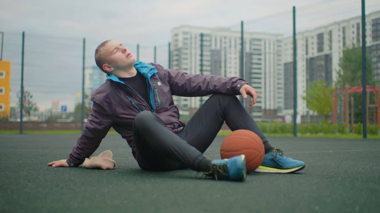 young man sitting on basketball court, clutching ball and adjusting jacket, pausing after drill, massaging neck and taking deep breaths, skyline of apartment towers beyond fenced court, overcast sky