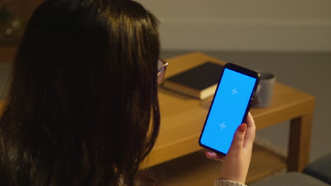 vista trasera de una mujer joven pasando la noche en casa sentada en el sofá mirando la pantalla azul del teléfono móvil 3